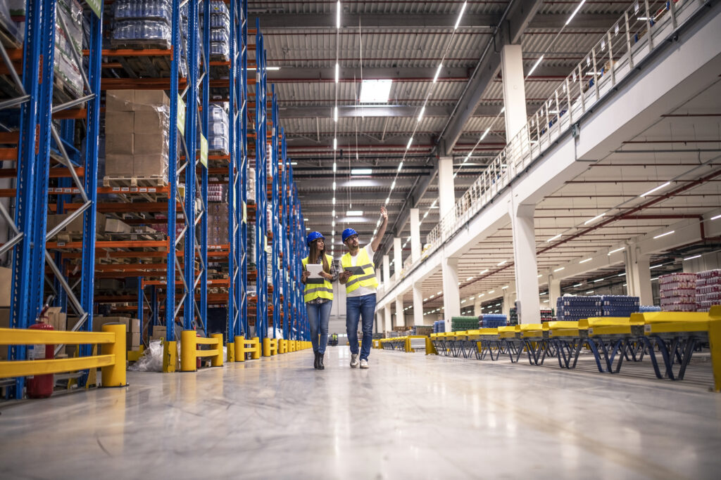 Distribution warehouse interior with workers wearing hardhats and reflective jackets walking in storage area - signs your warehouse needs a WMS