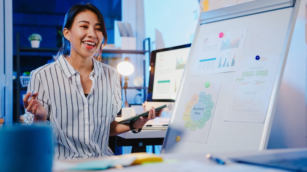 Asia businesswoman social distancing in new normal for virus prevention looking at camera presentation to colleague about plan in video call while work in office night. Lifestyle after coronavirus.